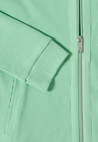 Lysegrøn jakke i tekstureret stof med en sølvfarvet lynlås og en opklappet ærmekant, som fremhæver strikkemønsteret og lommedetaljen.