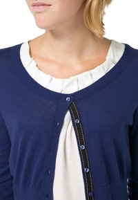 Námořnická modrá kardigán s kulatým výstřihem, zdobený řadou modrých knoflíků a dekorativním černým lemem s bílým šitím. Nosený přes bílou volánkovou halenku.