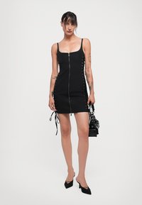 Black bodycon dress with a front zipper, adjustable side gathers, and lace-up detailing. Smooth fabric and slim fit, paired with pointed heels.