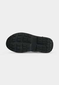 Semelle d'une chaussure noire avec un motif de bande de roulement rectangulaire et carré texturé pour adhérence, présentée sur un fond gris clair uni.