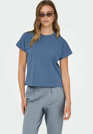 Giovane donna con maglietta a maniche corte blu, pantaloni grigi a vita alta e occhiali da sole marroni a forma di occhio di gatto, in piedi con postura rilassata.