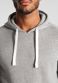 Grå huvtröja i mjukt material med en framficka i kängurustil, vita snörningar och en avslappnad halsringning. Slät textur med minimal detaljering.