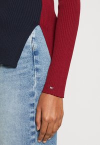 Top a maniche lunghe a coste con blocchi di colore blu navy e rosso; presenta una piccola accentatura con il marchio sul polsino. Abbinato a jeans blu chiaro.