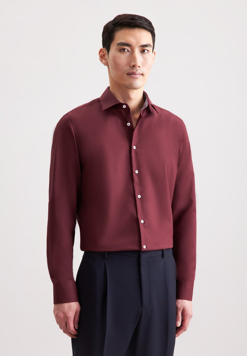 Camicia a maniche lunghe color bordeaux realizzata in tessuto liscio, con un design aderente, bottoni bianchi e un colletto con un motivo discreto. Abbinata a pantaloni scuri.