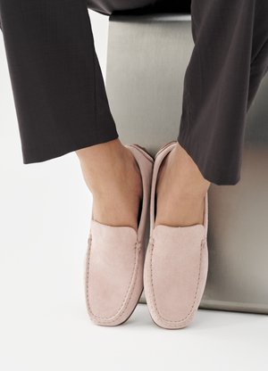 Άτομο που φοράει σουέντ μοκασίνια σε ανοιχτό ροζ χρώμα και σκούρο παντελόνι, με τα πόδια να ακουμπούν σε μεταλλικό ορθογώνιο μπλοκ σε λευκό φόντο.