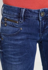 Primer plano de jeans de mezclilla azul con bolsillo con cremallera y botón, usados con una blusa color coral.