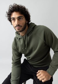 Olive rohelise kapuutsiga sulejope, millel on stringid, soonilised varrukad ja tekstureeritud pind. Kanda mustade pükstega heledate hallide taustal.