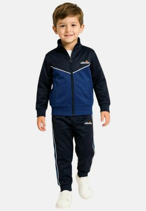 Jeune garçon souriant portant un survêtement ellesse bleu marine et noir avec des baskets blanches, debout devant un fond uni.