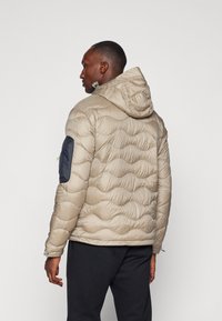 Beige puffer jacket with quilted texture and hood. Features black accents on the sleeves and a fitted silhouette. Lightweight and insulated.