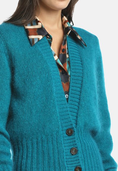 Tiefergrün gestrickte Strickjacke mit V-Ausschnitt, geripptem Saum und fünf braunen Knöpfen. Über einem gemusterten Hemd mit Kragen getragen.
