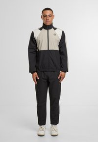 Giacca a vento nera e beige con colletto alto, polsini elasticizzati e pantaloni abbinati. Presenta un logo sul petto e dettagli laterali.