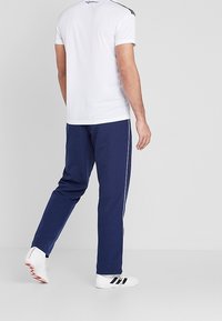 Man som står, iklädd en vit kortärmad skjorta, marinblå träningsoverallsbyxor med vita ränder och vita sneakers med svarta och röda detaljer.