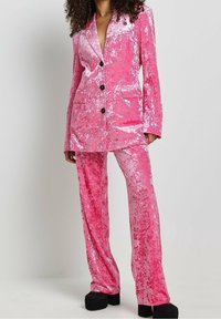 Rosa Samtanzug mit einem einreihigen Blazer, zwei Taschen, drei schwarzen Knöpfen und passenden Schlaghosen. Glatte Textur.