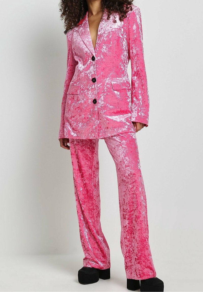 Rosa Samtanzug mit einem einreihigen Blazer, zwei Taschen, drei schwarzen Knöpfen und passenden Schlaghosen. Glatte Textur.
