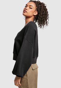 Ung kvinne med krøllete hestekur og svart oversized genser og beige bukser, stående i profil mot en ensfarget bakgrunn.