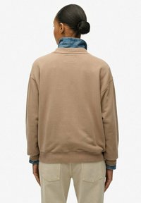 Beżowa bluza z luźnym krojem, wykonana z miękkiego materiału, z dżinsowym kołnierzem i ściągaczami, przedstawiona z tyłu w połączeniu z jasnymi spodniami.