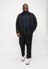 Svart zip-jakke over en marineblå polo, kombinert med løse svarte bukser og hvite sneakers med svarte og blå detaljer. Glatt tekstur totalt.