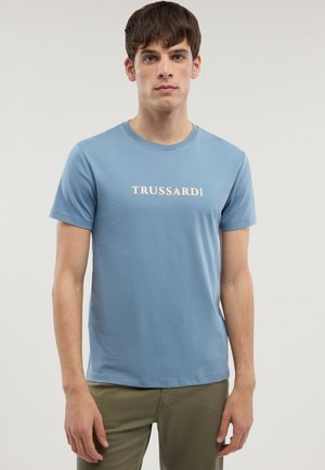 Jonge man met een lichtblauw T-shirt met de tekst "TRUSSARDI", gecombineerd met olijfgroene broek, staand tegen een effen lichte achtergrond.