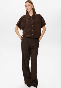 Woman standing with hands in pockets wearing dark brown short-sleeve button-up shirt and matching wide-leg pants with pointed shoes.