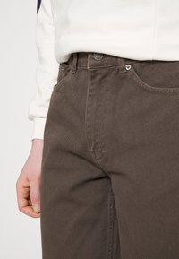 Ruskeat puuvillaiset corduroy-housut suoraa leikkausta, joissa on nappikiinnitys ja taskut sivuilla, esittäen teksturoitua kangasta ja hienovaraisia ompelutietoja.