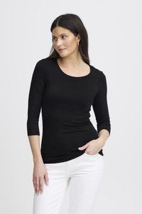 Donna con lunghi capelli scuri che indossa una maglia nera a maniche tre quarti e pantaloni bianchi, in piedi con una mano in tasca, guarda di lato.
