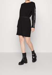 Svart långärmad klänning med en samlad midja, prydd med ett metallicbälte och kontrasterande vit märketext på ärmarna. Modell som bär svarta klumpiga stövlar.