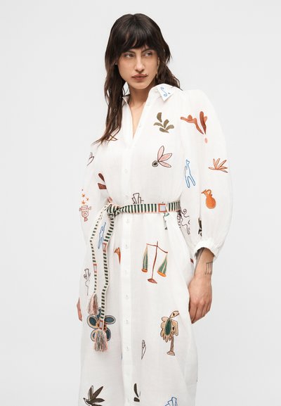 Γυναίκα που φοράει λευκό φόρεμα με κουμπιά, διακοσμημένο με πολύχρωμα κεντημένα αφηρημένα σχήματα και ριγωτή ζώνη δεμένη στη μέση, στέκεται μπροστά σε απλό φόντο.