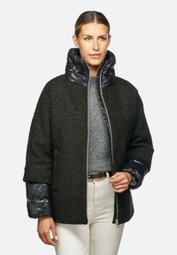 Cappotto nero con texture, maniche e colletto imbottiti, zip dorata e maglione grigio sotto. Il modello si trova in piedi con le mani ai lati.