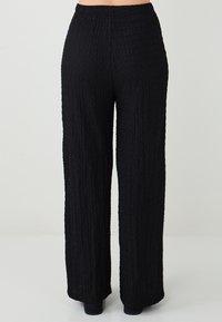 Persoon in hoge taille, met wijde zwarte textuurbroek, staande op een witte vloer tegen een effen lichte achtergrond.