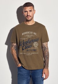 Olivově zelené bavlněné tri čko s modrým grafickým textem a logem. Kulatý výstřih, krátké rukávy, volnější střih, kombinované s modrými džíny.