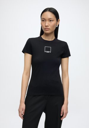 Kobieta z prostymi czarnymi włosami, ubrana w dopasowany czarny T-shirt z małym białym kwadratowym logo oraz czarne spodnie na jednolitym tle.