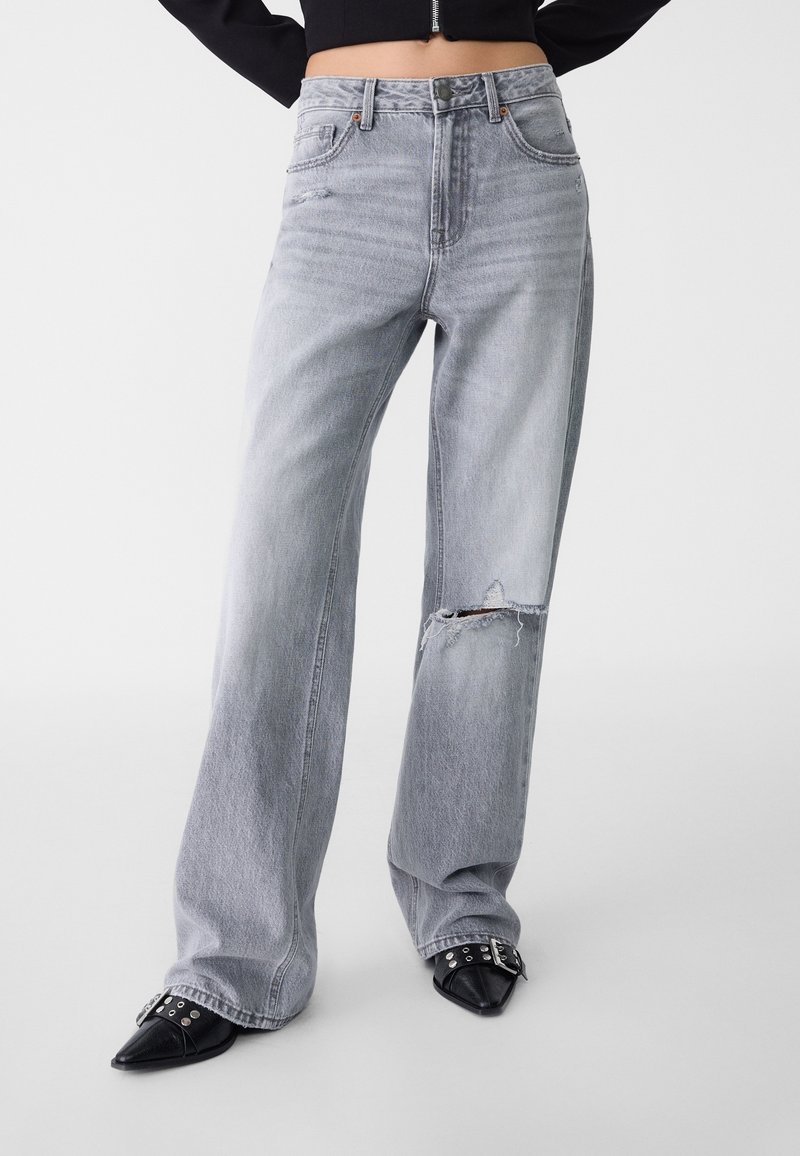 Personne portant un jean gris clair à jambes larges avec un genou déchiré et des chaussures pointues noires à boucles sur fond blanc.