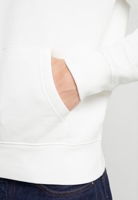 Sudadera blanca con un bolsillo tipo canguro, puños acanalados y dobladillo. La tela parece suave y ligeramente texturizada. Se ven unos jeans de mezclilla oscura.