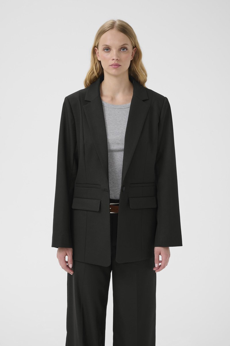 Fekete szabott blézer, hullámos gallérral, két első zsebbel és egysoros gombolással. Szürke felső fölött viselve, az anyag simának tűnik.