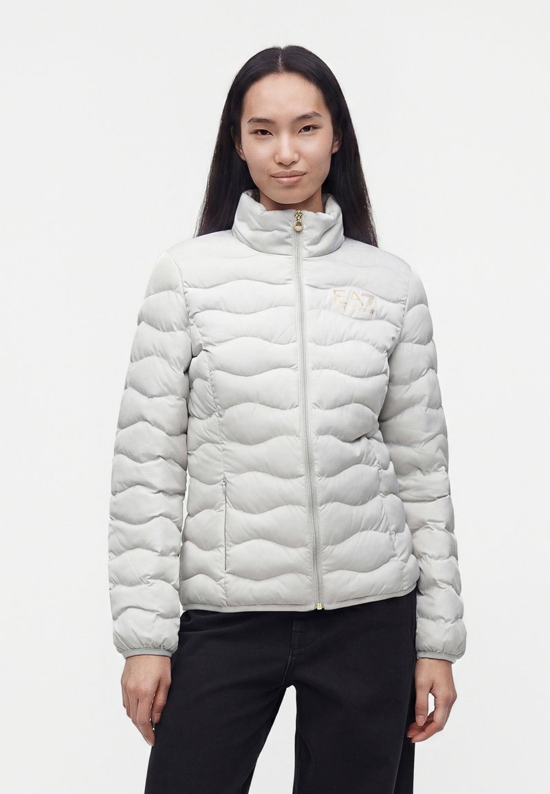 Jeune femme aux longs cheveux foncés portant une doudoune matelassée gris clair et un pantalon noir, debout devant un fond blanc uni.
