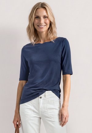 Kvinde med skulderlangt blondt hår smilende, iført en mørkeblå top med korte ærmer og hvide bukser, der holder en brun skuldertaske.
