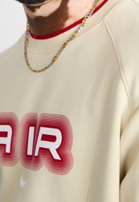 Sudadera beige con cuello redondo acanalado y acentos rojos, que presenta la audaz inscripción "AIR" en rojo y blanco. Cadena de collar visible.