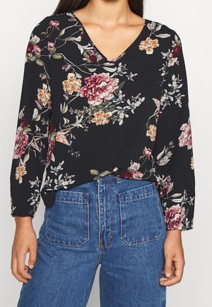 Blouse noire col V avec imprimé floral rose, orange et blanc, associée à un jean bleu taille haute avec poches plaquées à l'avant.