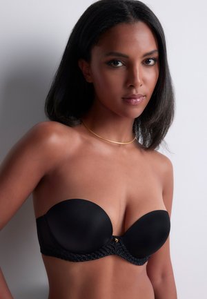 Femme aux cheveux noirs mi-longs portant un soutien-gorge noir sans bretelles et un fin collier en or, sur un fond clair uni.