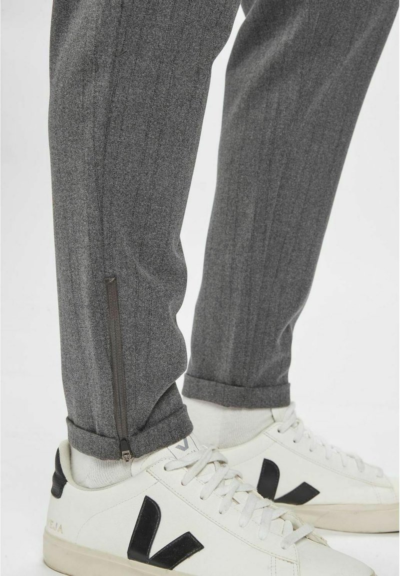 Untere Beine in grauen Hosen mit Reißverschlüssen an den Knöcheln und weißen Sneakern mit schwarzen "V"-Logos, kombiniert mit weißen Socken vor einem weißen Hintergrund.