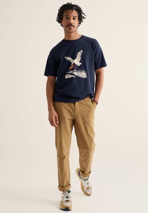 Man loopt, draagt een marineblauw t-shirt met vogelafbeelding, kaki broek opgerold bij de enkels en multicolor sneakers, tegen een lichte achtergrond.