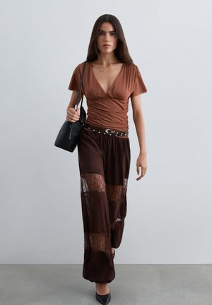 Mujer caminando con una blusa de cuello en V color óxido, pantalones marrón oscuro transparentes con paneles de encaje, tacones negros, cinturón con tachuelas y lleva un bolso de mano negro.