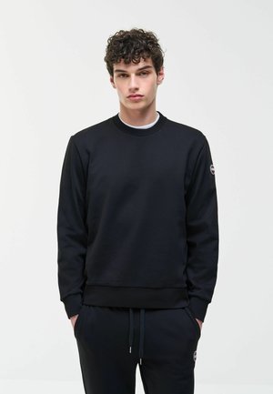 Noor mees lokkis juustega, kannab musta dressipluusi ja sobivaid musti dressipükse, käed taskutes, ühtlase taustaga.