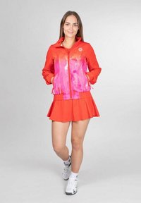 Rode sportjacket met een roze verfachtige print, voorzien van een ritssluiting aan de voorkant en zakken, gecombineerd met een bijpassende rode geplooide rok.