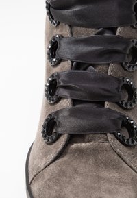 Stivale alla caviglia in suede grigio con lacci in satin nero attraverso occhielli decorativi neri accentuati da cristalli, evidenziando la texture e i dettagli.
