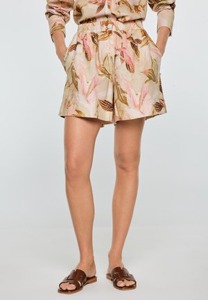 Kobieta w beżowych szortach w kwiaty z kieszeniami i dopasowanym długim rękawem, w połączeniu z brązowymi sandałami wsuwanymi.