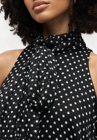 Κοντινό πλάνο ενός ατόμου που φορά ένα μαύρο τοπ χωρίς μανίκια με λευκές πουά και ένα ταιριαστό μαντήλι δεμένο γύρω από το λαιμό.