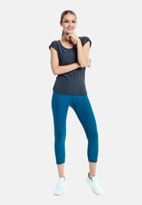 Gray short-sleeve top paired with teal leggings, both made from stretchy material. Model wears white athletic shoes, standing with arms crossed.
