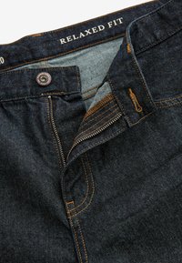 Tamsiai mėlynos denim medžiagos laisvi džinsai su kontrastine oranžine siūle, užtrauktuku ir žyma, kuriame užrašyta "LAISVAS KIRPIMAS" ant liemens.