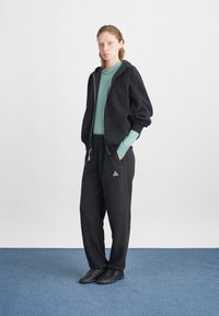 Μαύρο φούτερ με φερμουάρ και όγκωτους μανίκους, συνδυασμένο με ανοιχτό πράσινο μπλουζάκι μακρύ μανίκι και μαύρο παντελόνι φόρμας με ένα μικρό λογότυπο τριγώνου. Μαύρα υποδήματα.